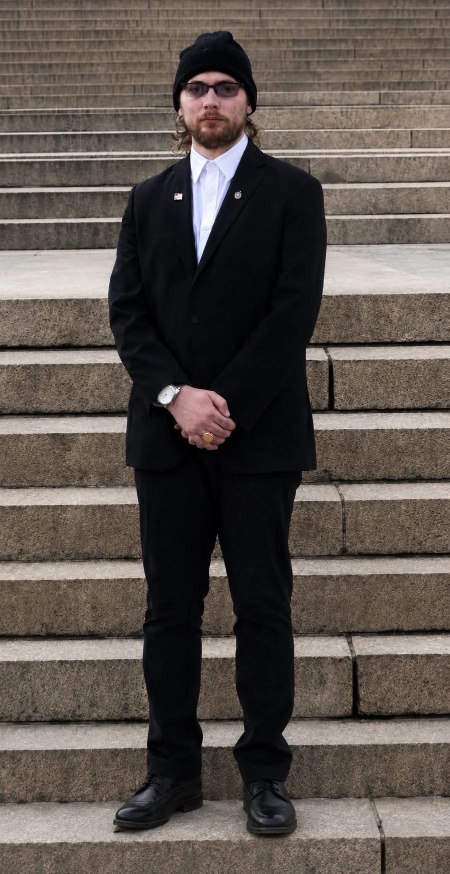 James Lacey standing on stone steps in a dark suit and black knit cap